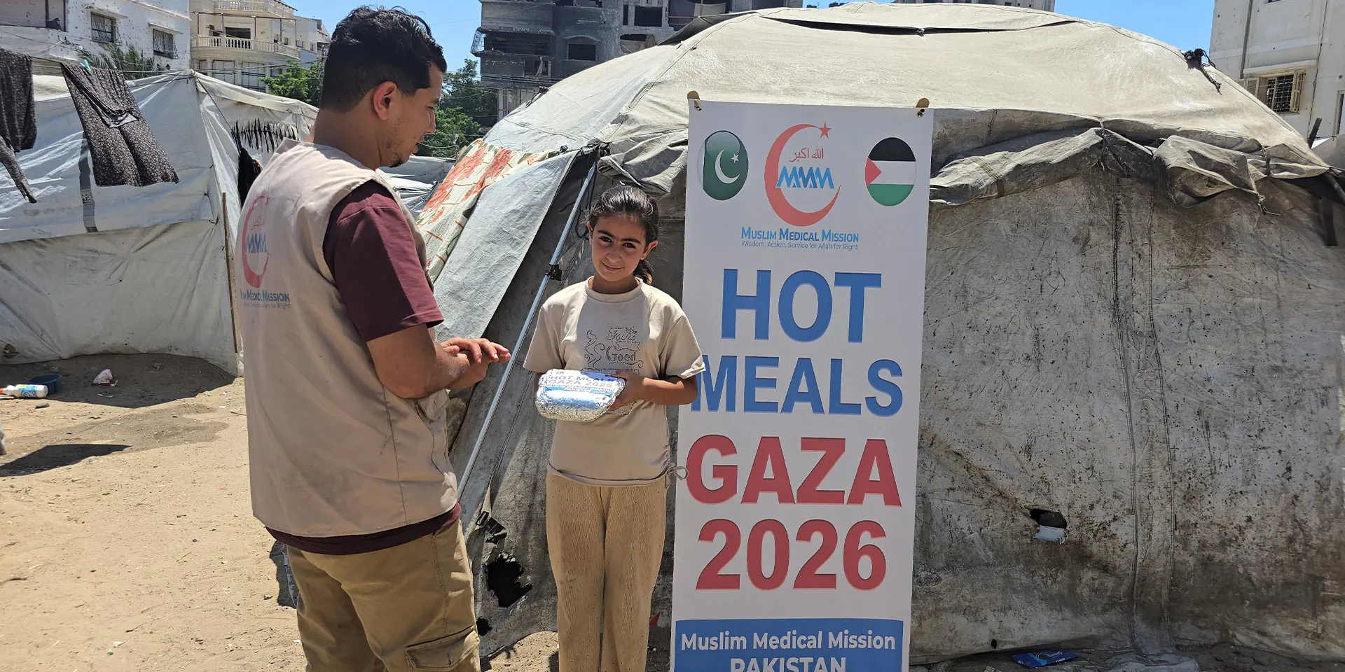 Muslim Medical Mission volunteer handing a hot meal to a child beside a Hot Meals Gaza 2026 banner at a tent camp in Gaza